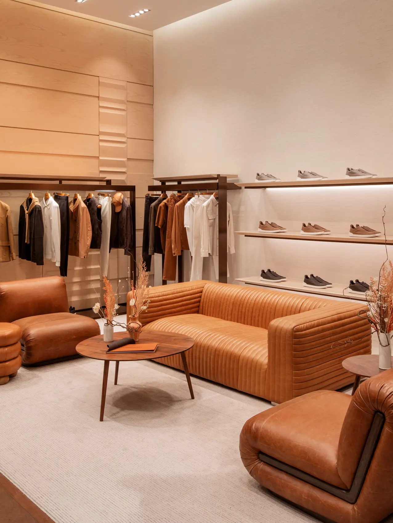 Image 8: Luxury men's boutique with a white carpet and leather armchairs. Shelves display ZEGNA sneakers, while jackets and coats are elegantly hung on a stand.