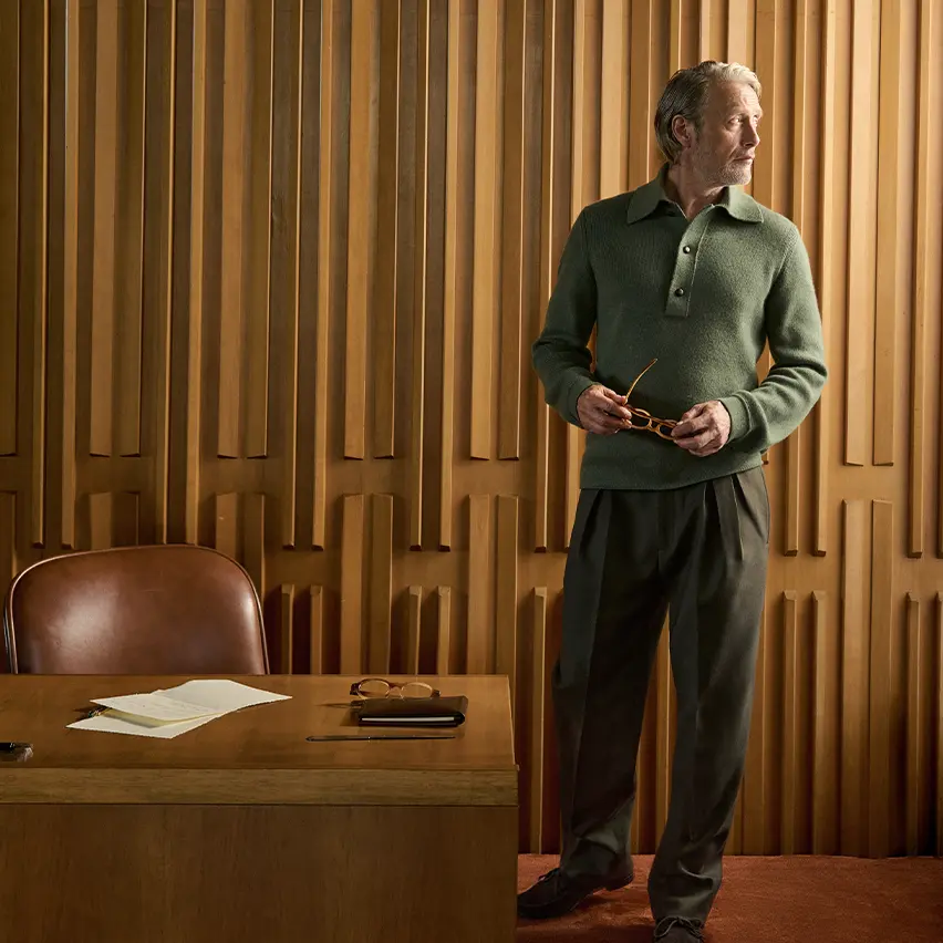 Mads Mikkelsen wearing a green ribbed knit polo with dark tailored trousers, holding his glasses in an elegant wood-paneled interior, from the ZEGNA Fall/Winter 2026 collection.