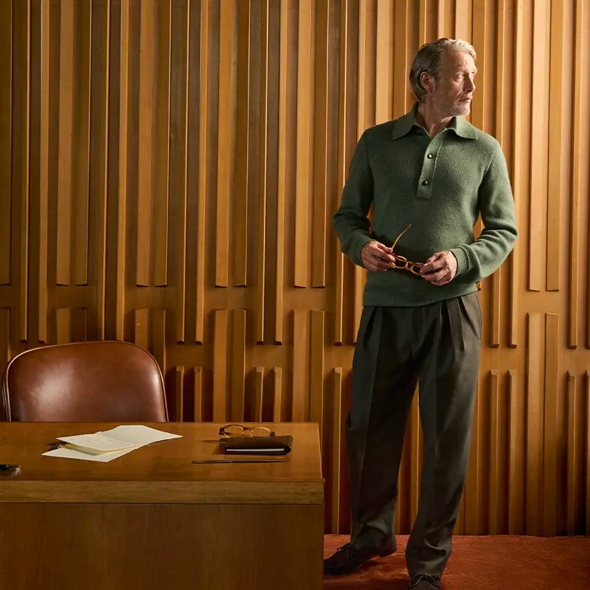 Mads Mikkelsen, vêtu d’un polo vert en maille côtelée et d’un pantalon d’esprit sartorial sombre de la collection Automne/Hiver 2026 de ZEGNA, tenant ses lunettes à la main dans un élégant intérieur lambrissé.