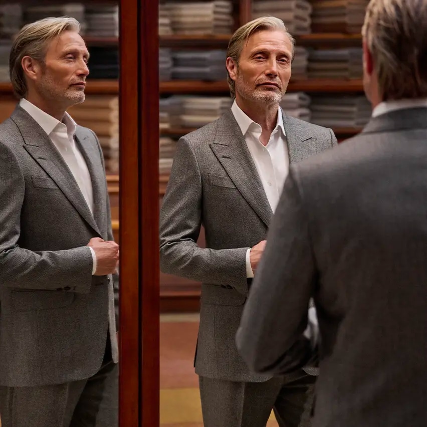 Mads Mikkelsen vêtu d’un élégant costume d’esprit sartorial gris et d’une chemise blanche de la collection Automne/Hiver 2026 de ZEGNA. Il apparaît en pied dans le triple miroir d’un atelier de tailleur.