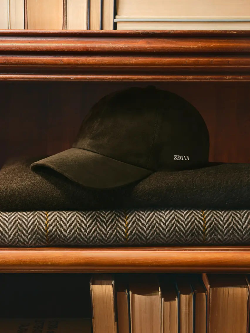 Eine Baseballkappe von ZEGNA, die auf gefalteten Schals in einem hölzernen Bücherregal liegt, aus der ZEGNA Herbst/Winterkollektion 2026.