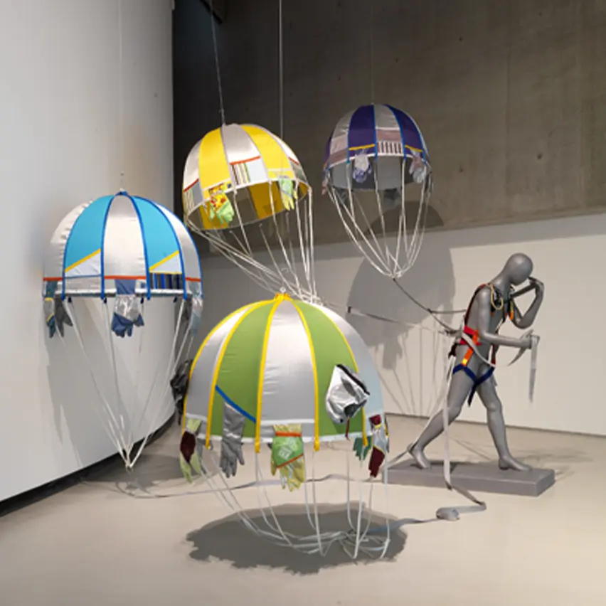 An installation featuring colorful parachutes suspended in the air, attached to a mannequin figure dressed in harness gear, representing the artwork 'Reconnaissance Man.'