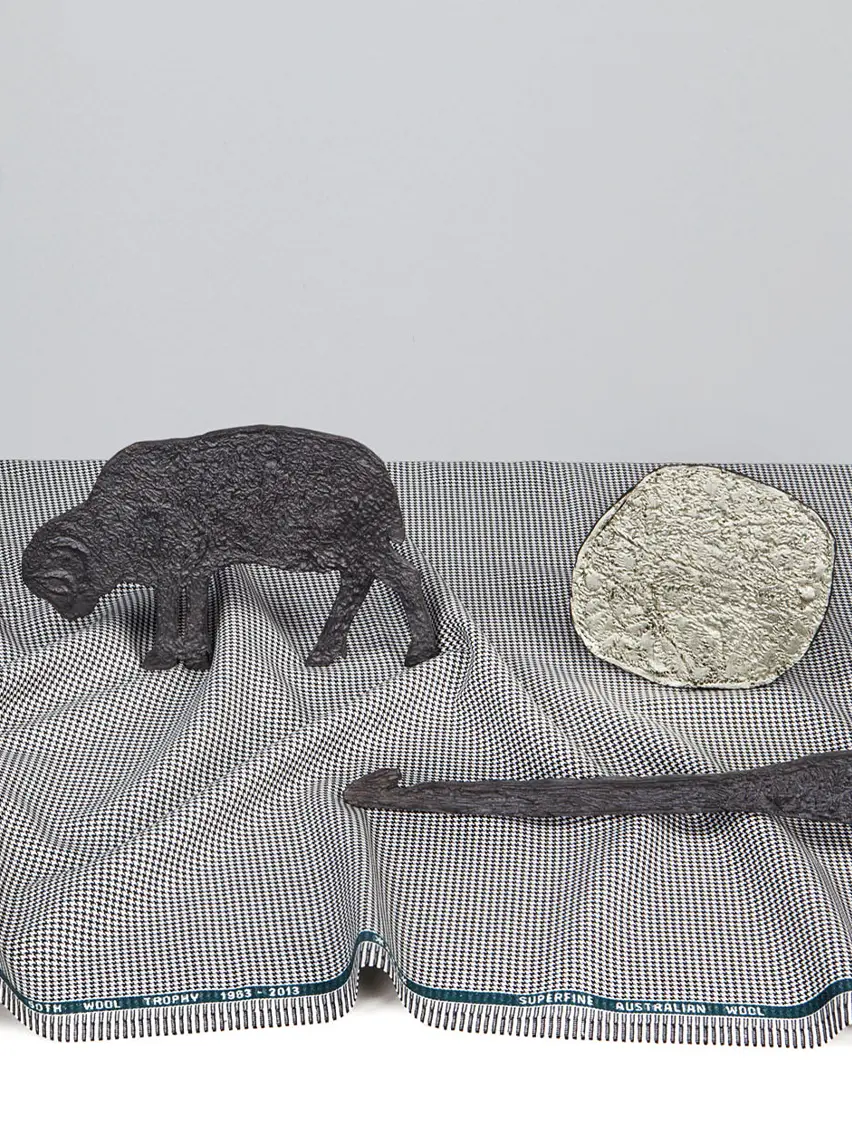 An artwork by Kiki Smith featuring a black figure of a sheep, a round textured object, and a long, dark shape on a patterned fabric background.
