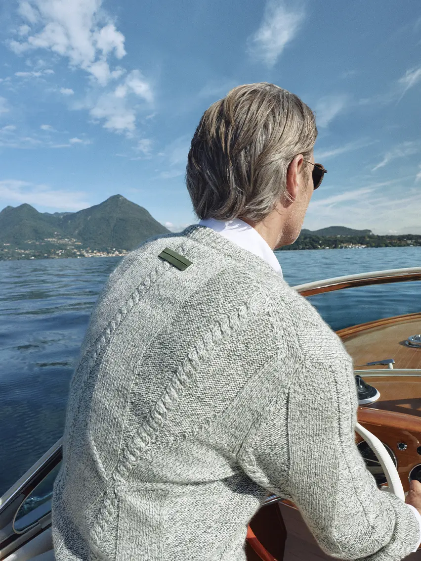 Close-up of Mads Mikkelsen on a boat by the lake wearing a grey V-neck sweater from the ZEGNA Spring Summer 2026 collection.