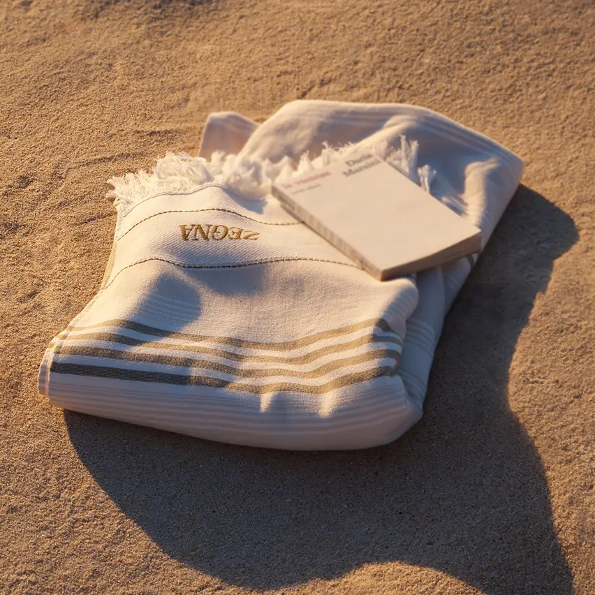 Beige cotton beach towel with a striped print, fringed trim on the short sides, and a prominently displayed ZEGNA logo, lying on the beach with a book on top.