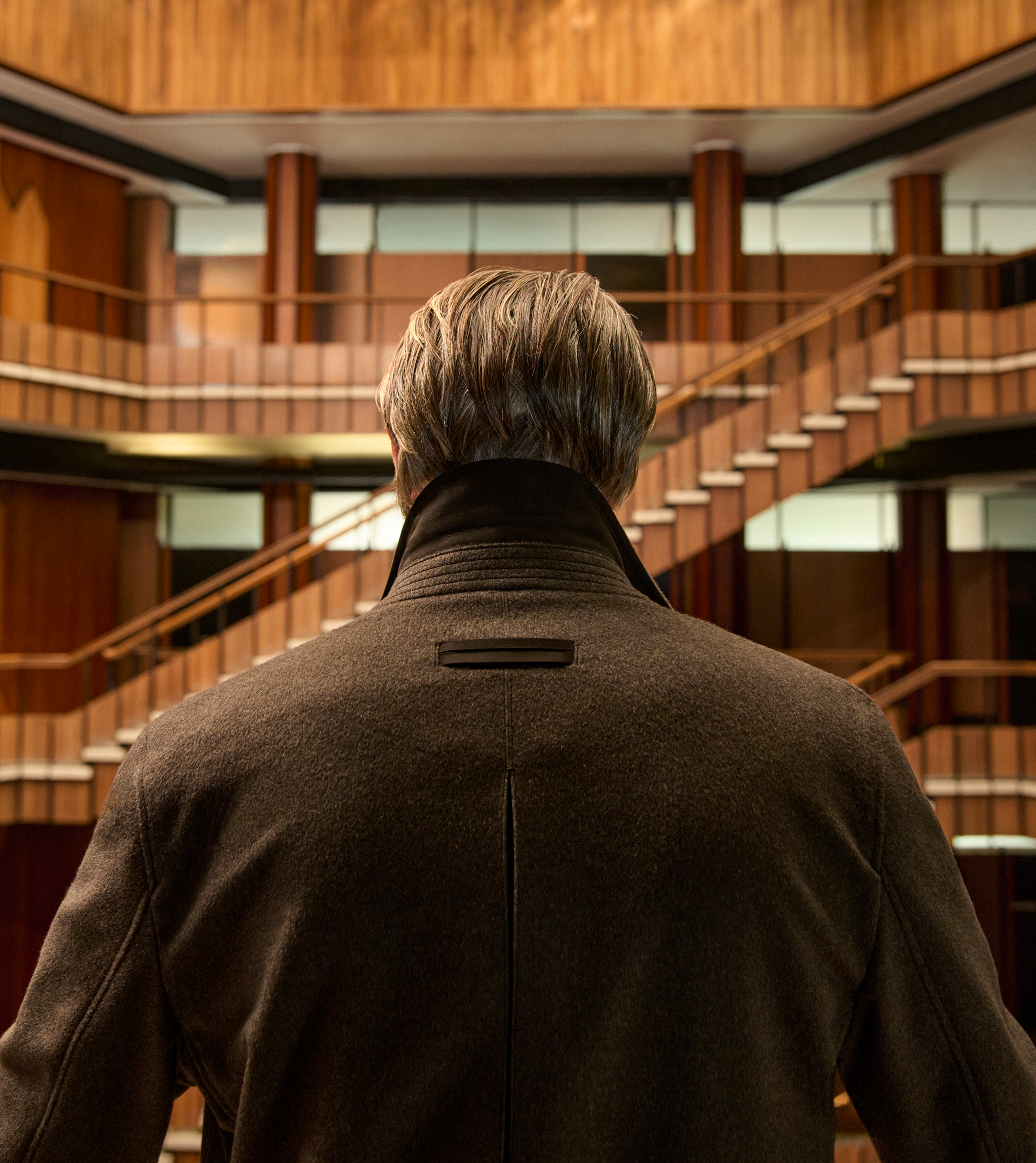 Mads Mikkelsen seen from behind in a dark brown wool coat, standing in a wood-paneled interior atrium, from the ZEGNA Fall/Winter 2026 collection.