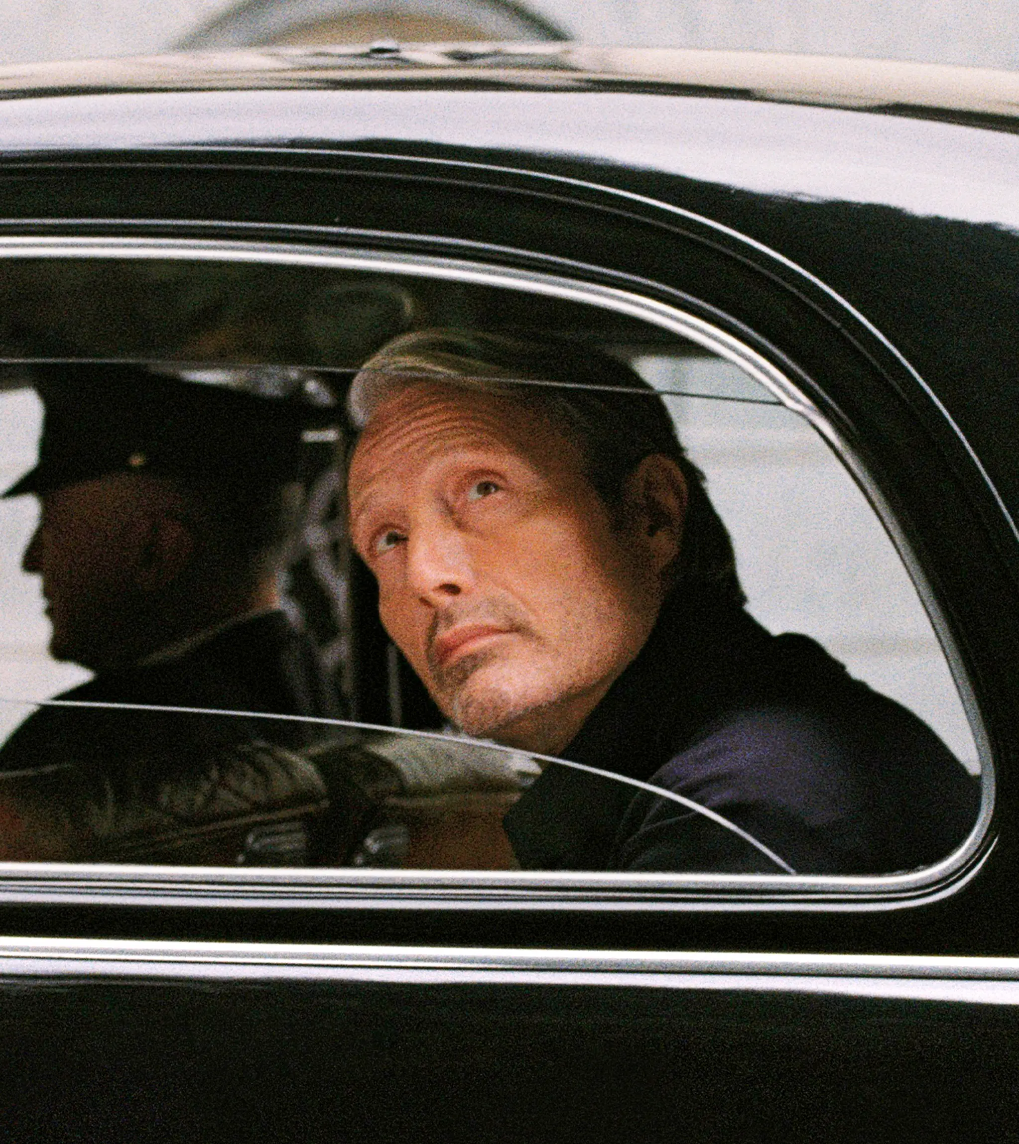 Mads Mikkelsen seated inside a classic black car, looking out through the side window.