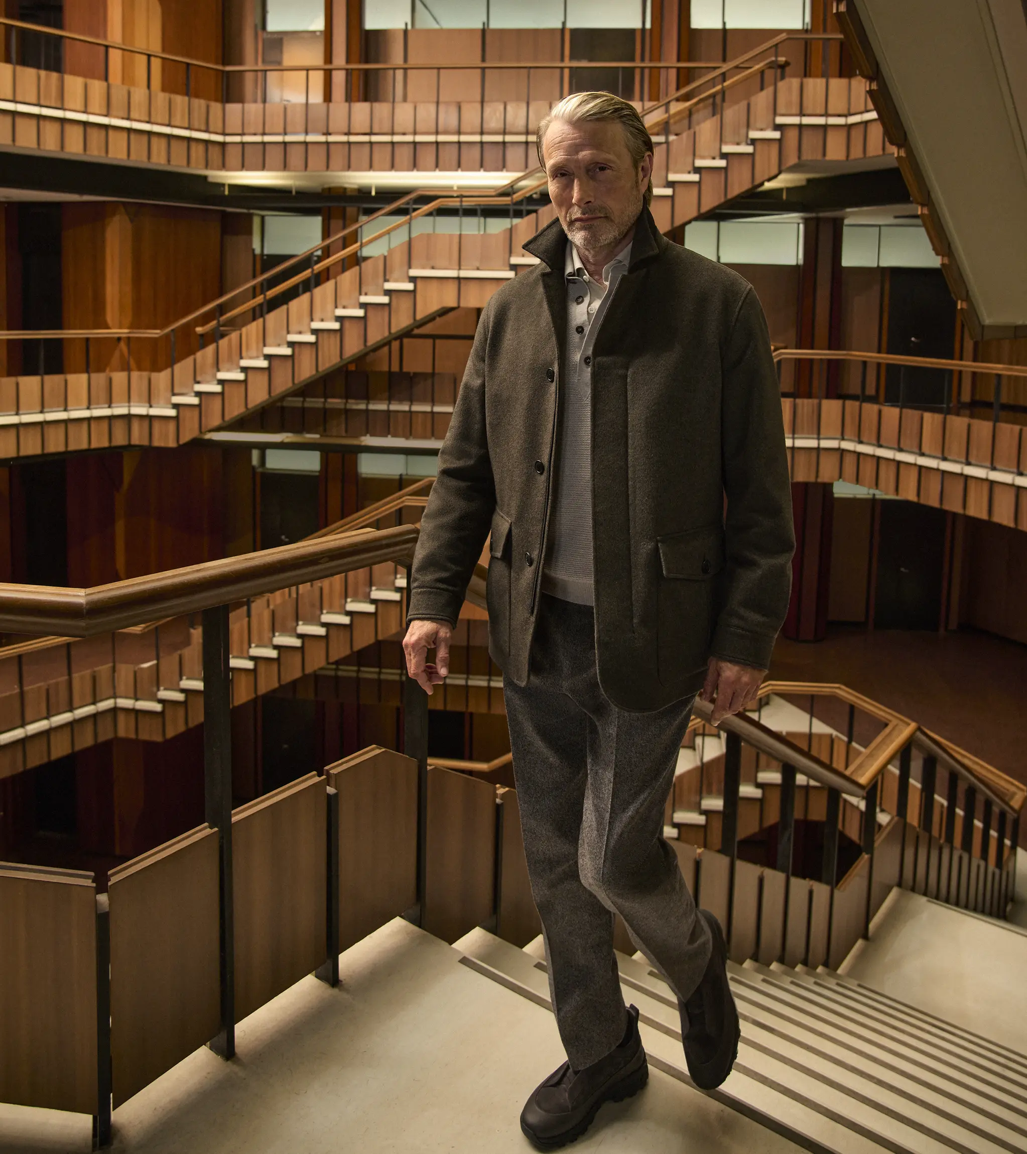 Mads Mikkelsen wearing a dark olive field jacket layered over a knit with tailored trousers and Triple Stitch shoes, on the staircase of a wood-paneled interior atrium, from the ZEGNA Fall/Winter 2026 collection.