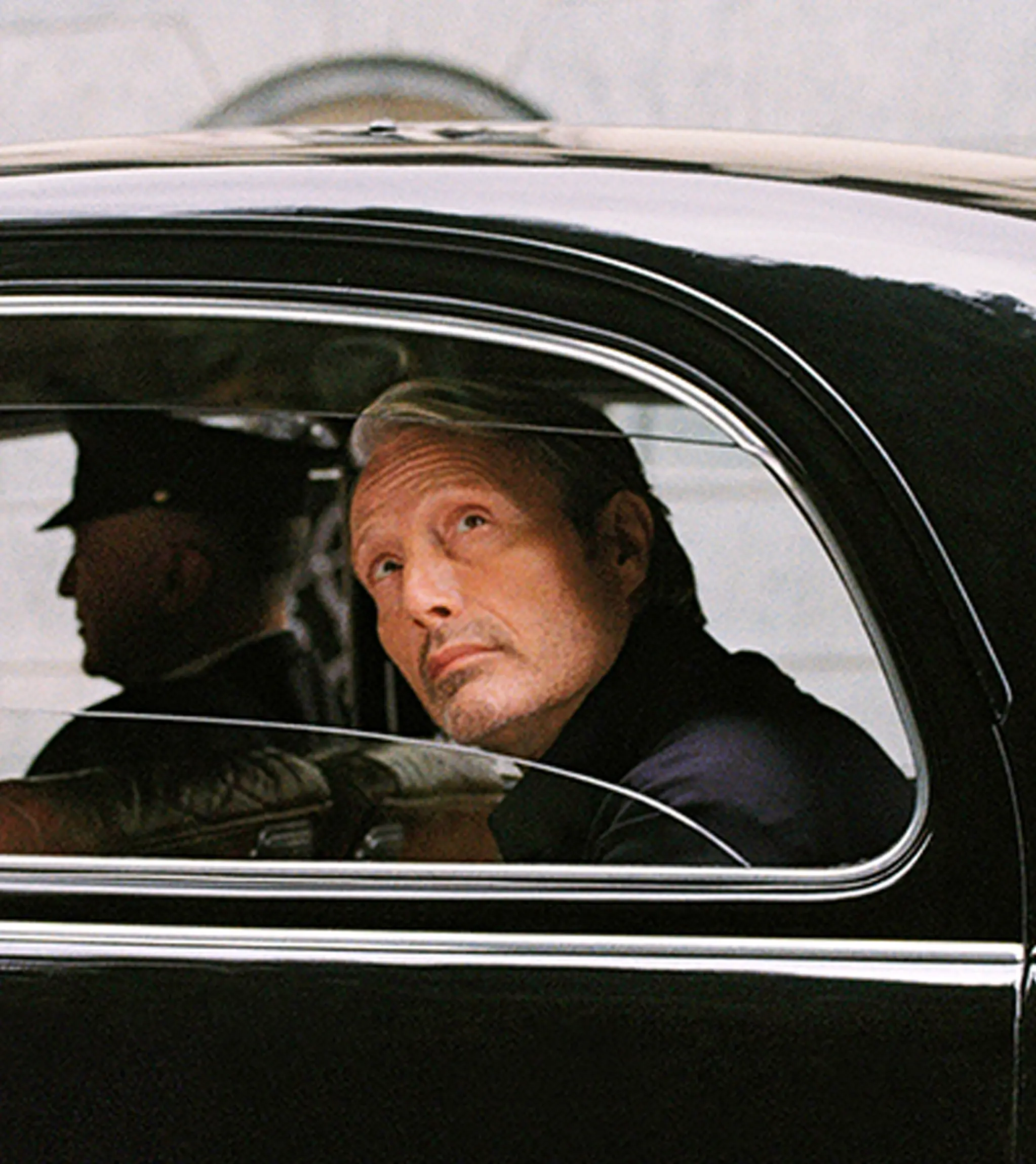 Mads Mikkelsen seated inside a classic black car, looking out through the side window.