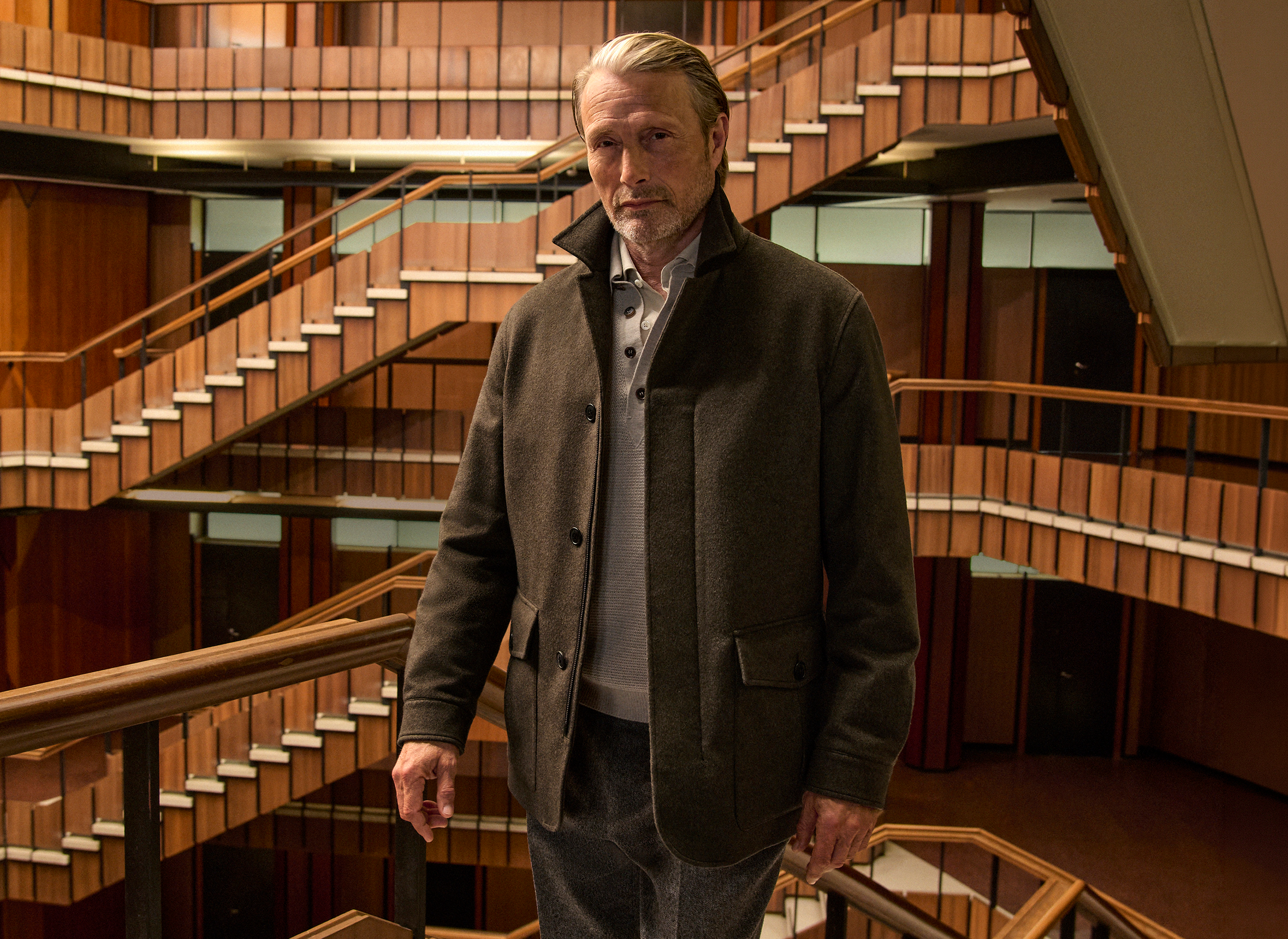 Mads Mikkelsen wearing a dark olive field jacket layered over a knit with tailored trousers and Triple Stitch shoes, on the staircase of a wood-paneled interior atrium, from the ZEGNA Fall/Winter 2026 collection.