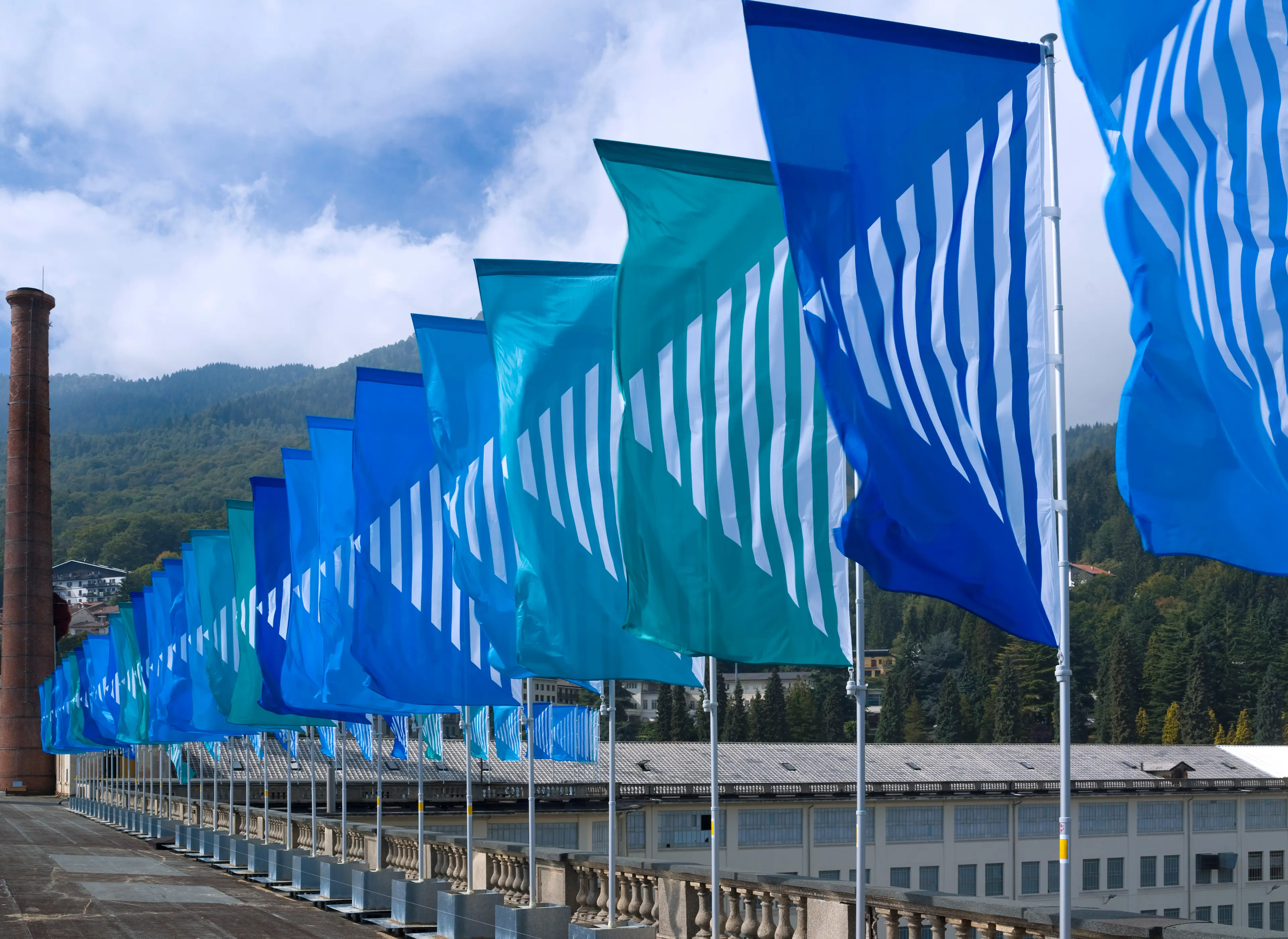 A view of Art Basel in Basel, featuring a historic building, a tall brick chimney, and vibrant blue flags lining a walkway.