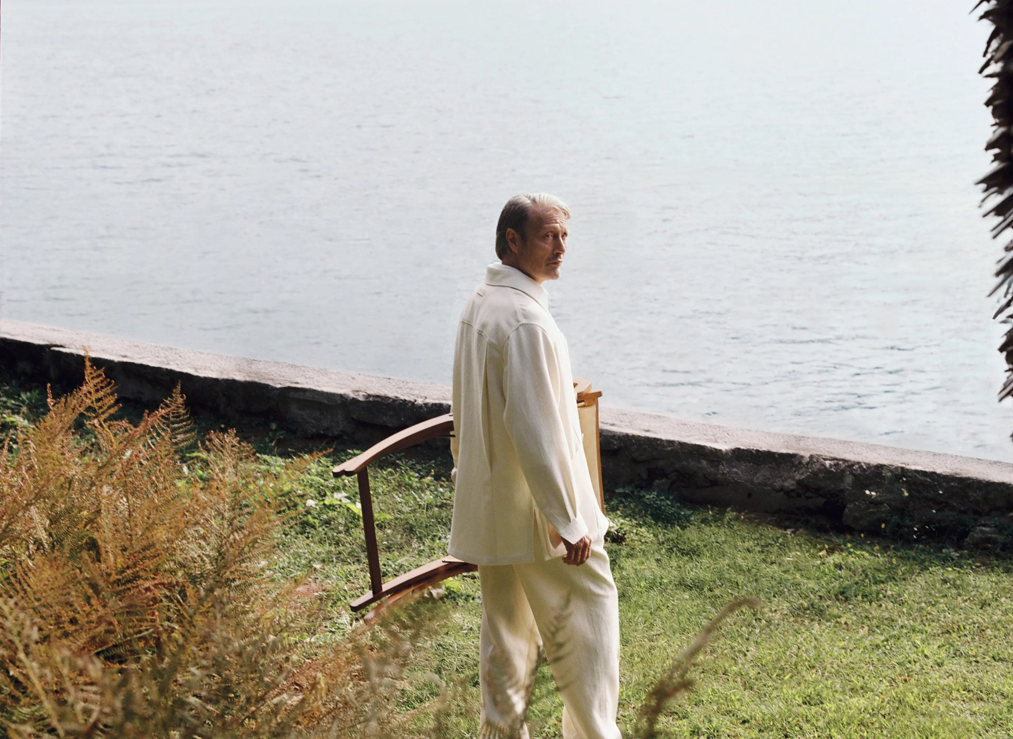 Mads Mikkelsen wearing a refined look from the ZEGNA Spring Summer 2026 collection in a lakeside garden setting.