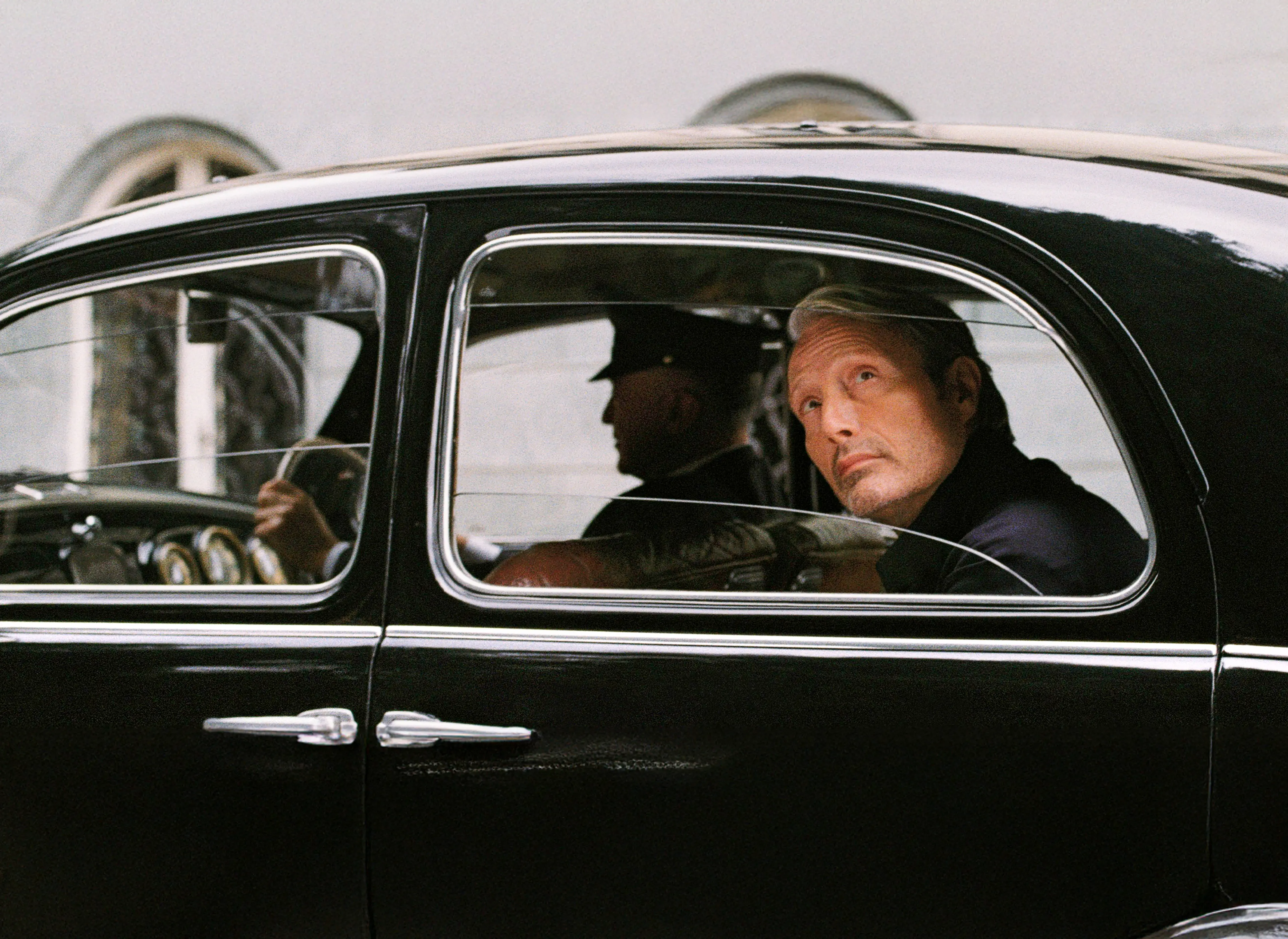 Mads Mikkelsen, seduto all’interno di un’automobile nera, osserva il paesaggio oltre il finestrino.