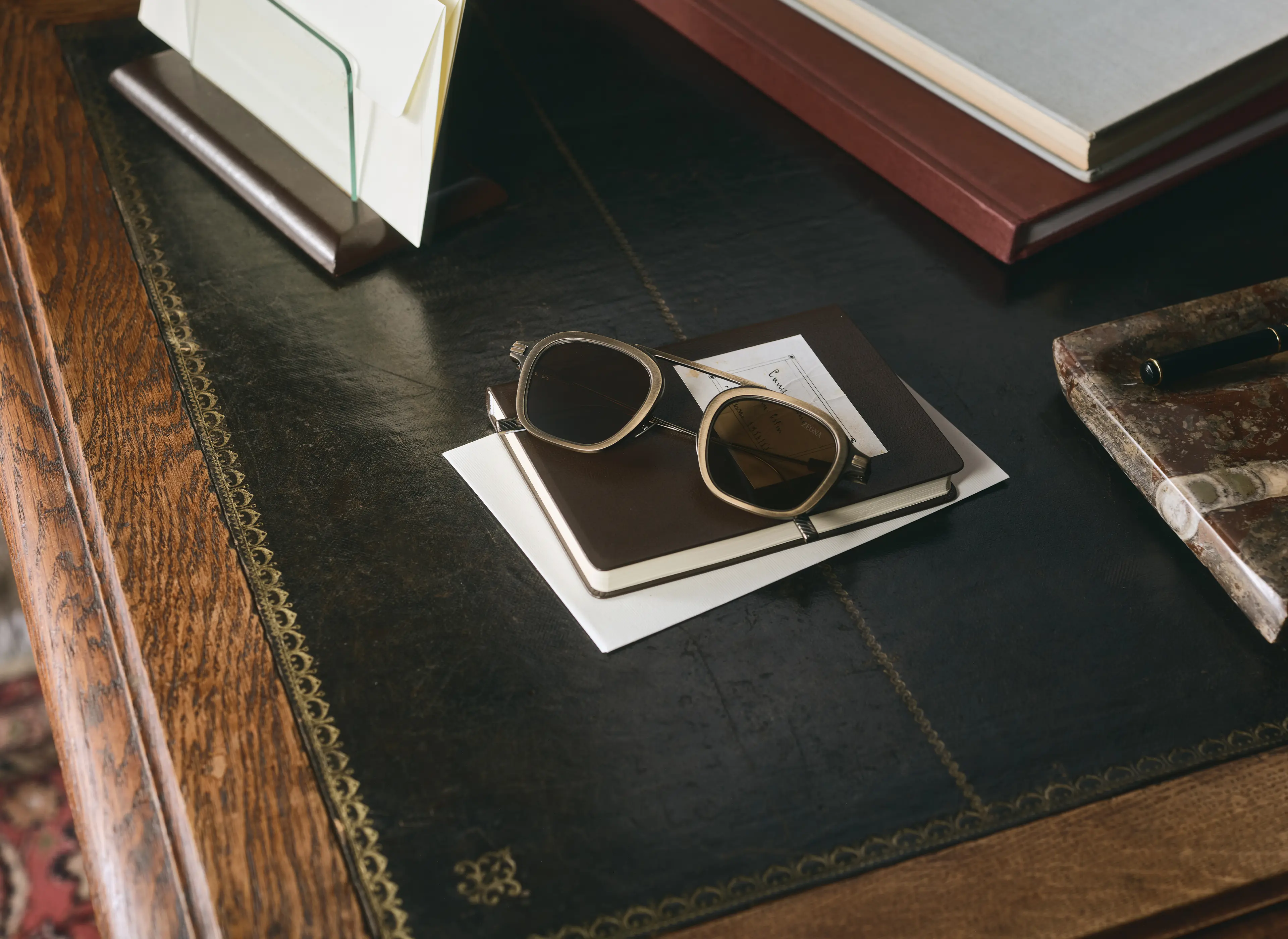 Eine ZEGNA Sonnenbrille aus Titan liegt auf einem Notebook, das auf einem eleganten Schreibtisch mit Lederoberfläche steht.