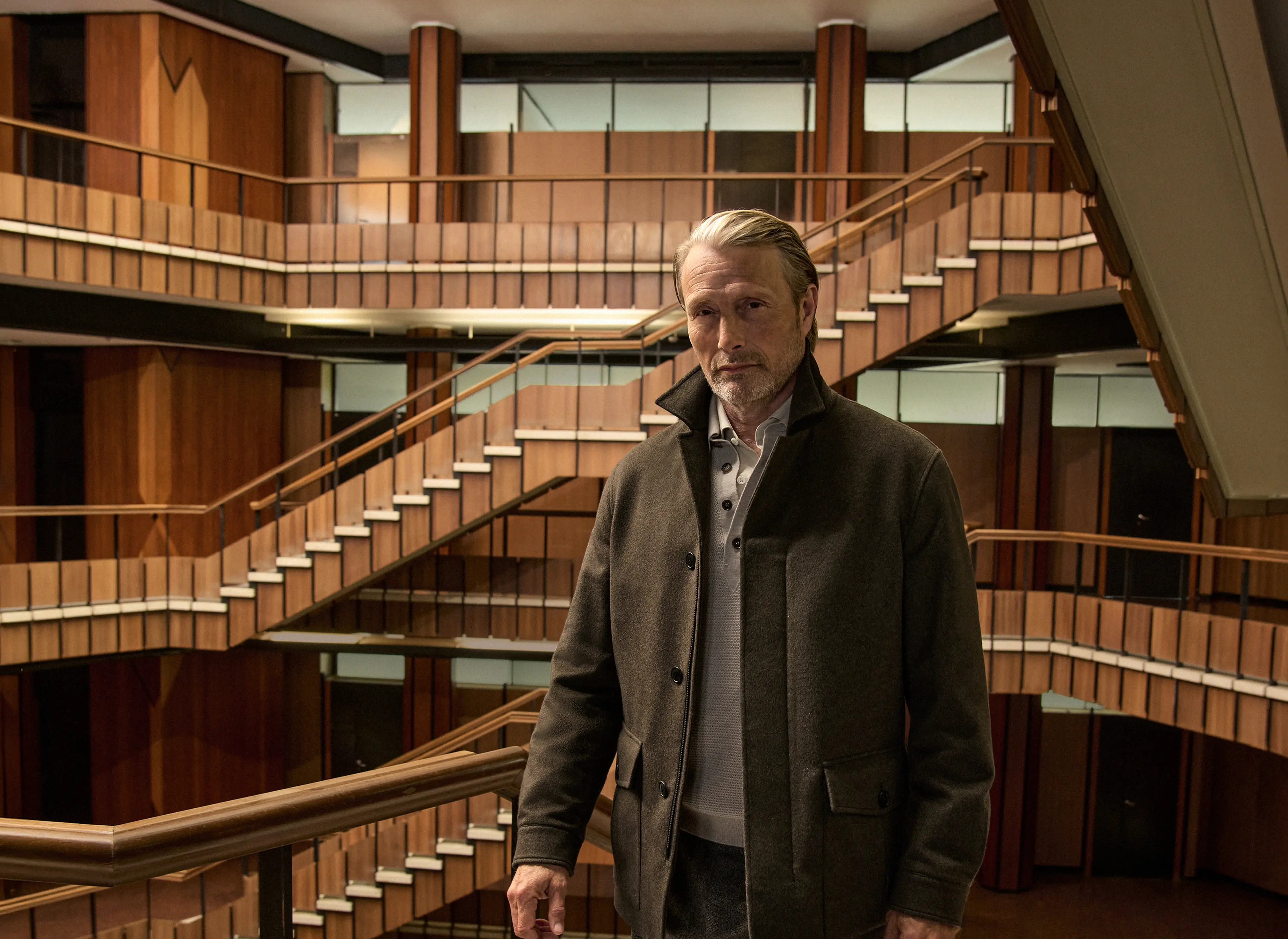 Mads Mikkelsen wearing a dark olive field jacket layered over a knit with tailored trousers and Triple Stitch shoes, on the staircase of a wood-paneled interior atrium, from the ZEGNA Fall/Winter 2026 collection.