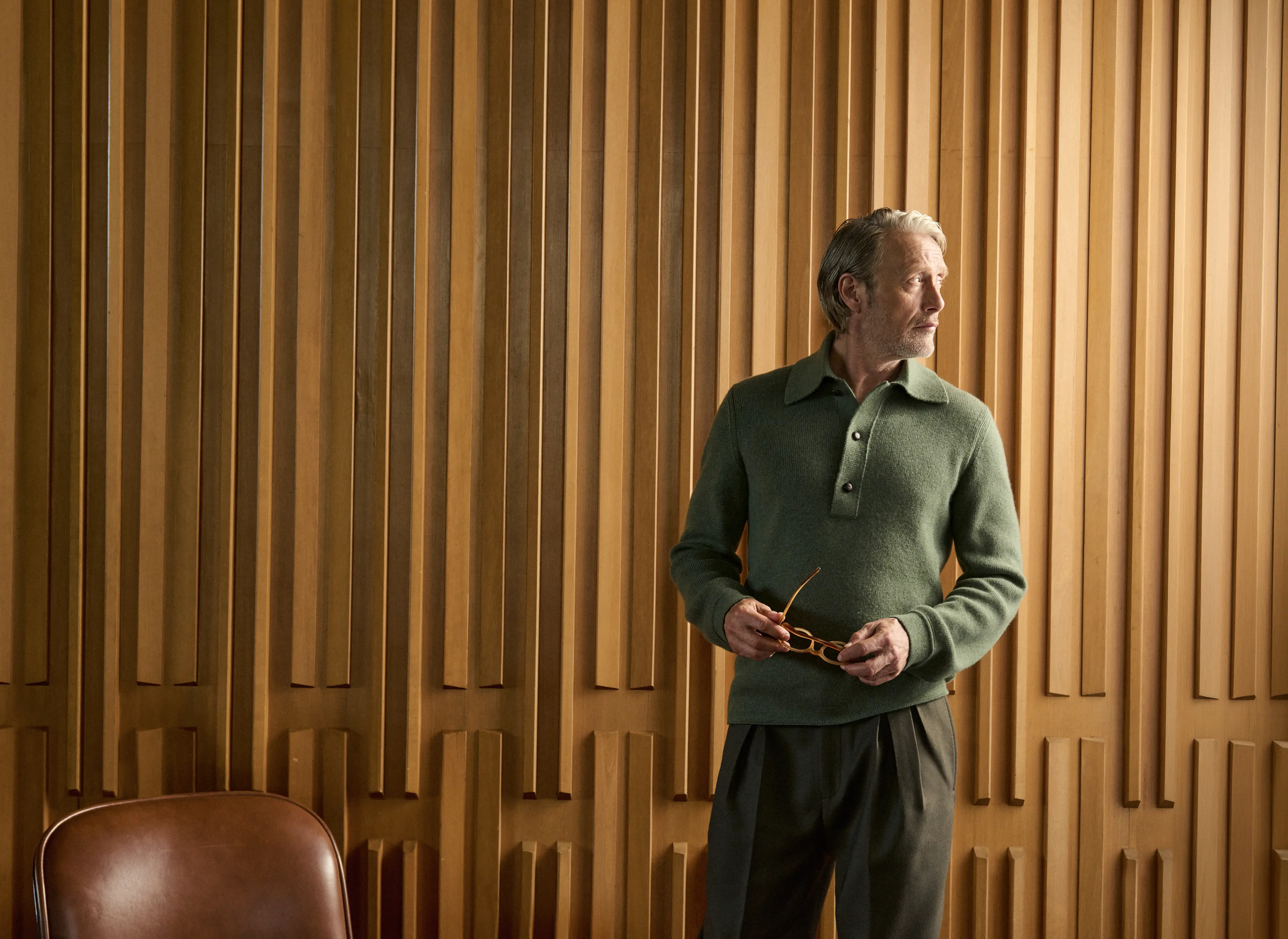Mads Mikkelsen wearing a green ribbed knit polo with dark tailored trousers, holding his glasses in an elegant wood-paneled interior, from the ZEGNA Fall/Winter 2026 collection.