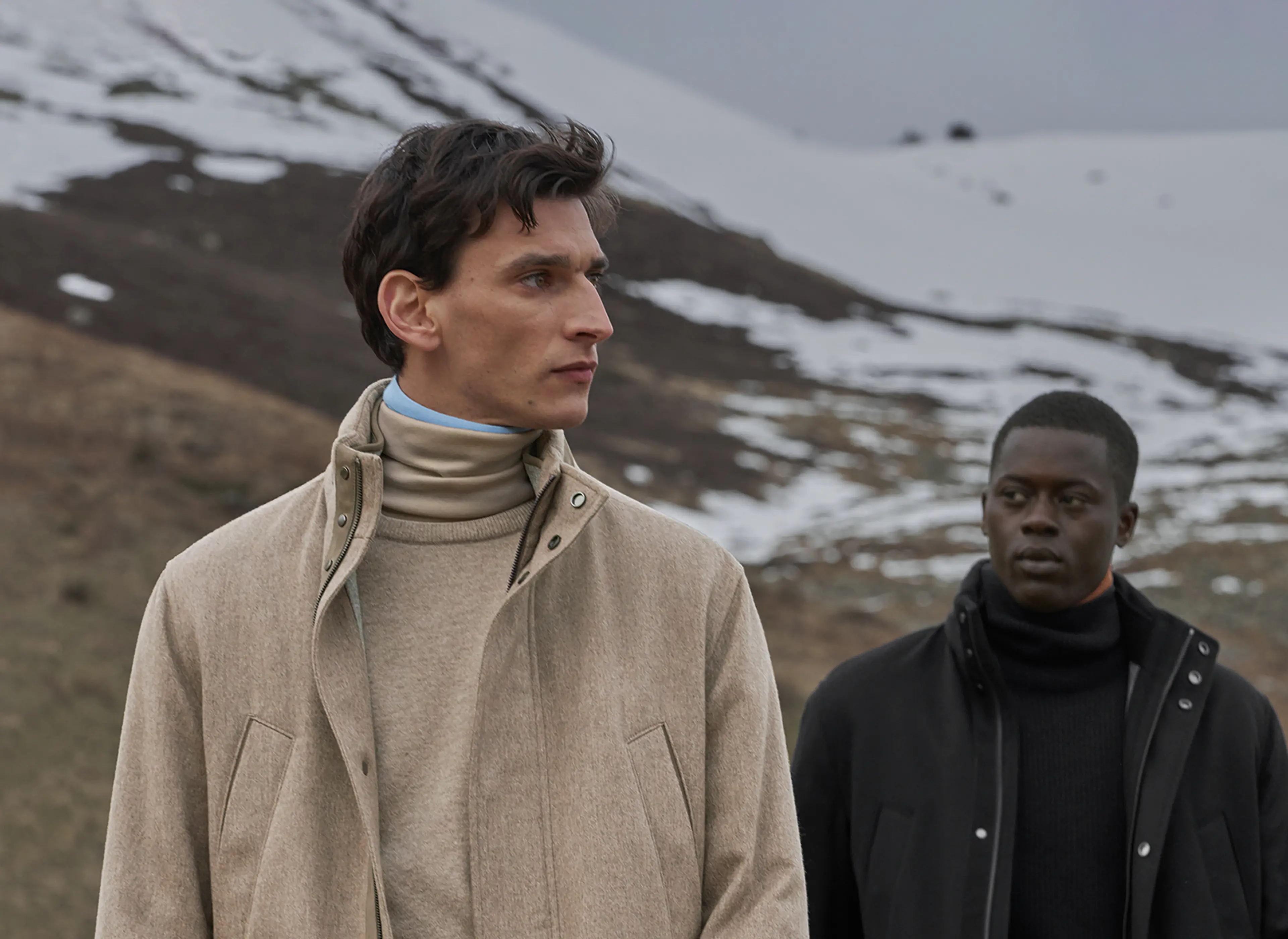 Dois homens com visuais monocromáticos em bege e preto, com calças de caxemira, golas altas e casacos acolchoados da nova coleção de outono/inverno da ZEGNA