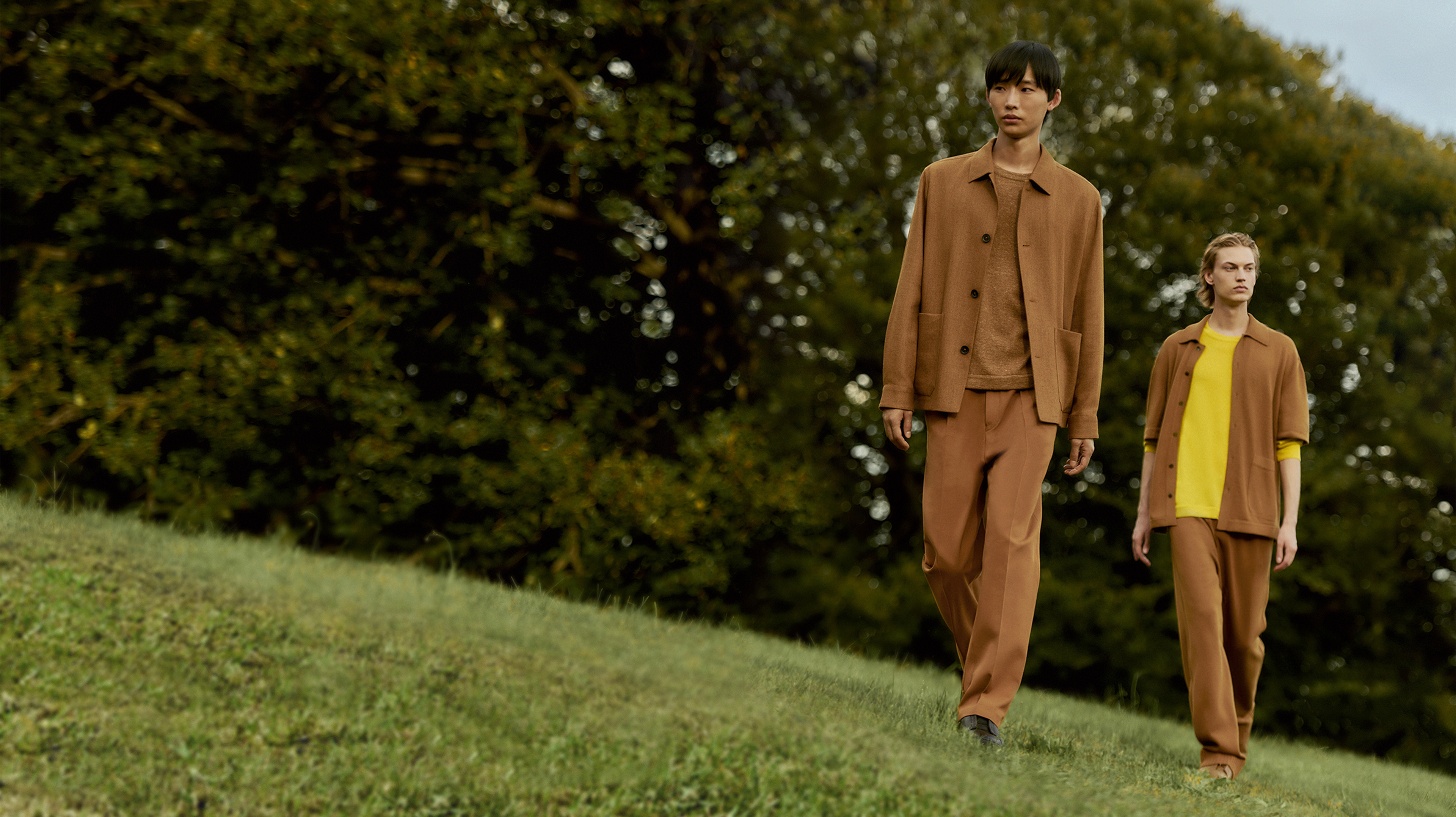 Two men walk on a lawn wearing outfits from ZEGNA's Oasi Cashmere Collection with brown overshirts and trousers.
