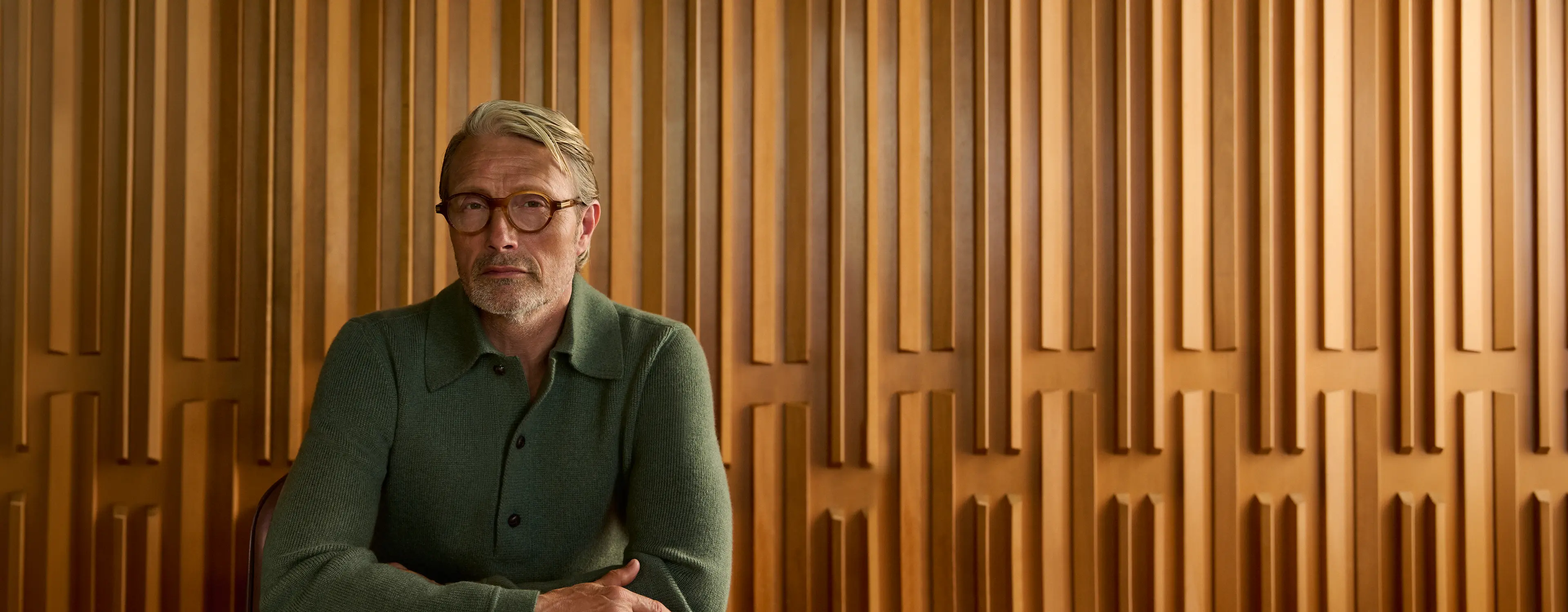 Mads Mikkelsen seated at a desk wearing a green ribbed knit polo with glasses, against a sculptural wood-paneled wall, from the ZEGNA Fall/Winter 2026 collection.