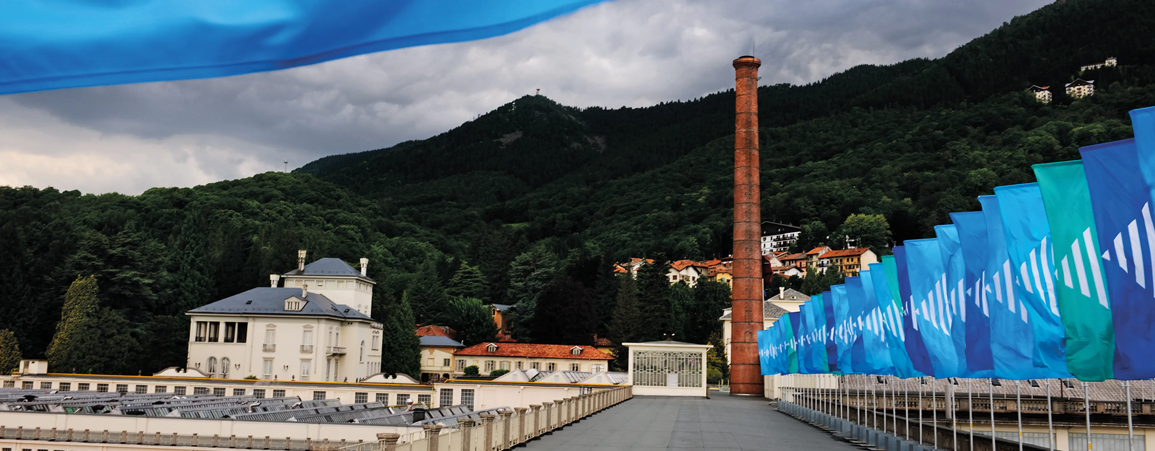 A view of Art Basel in Basel, featuring a historic building, a tall brick chimney, and vibrant blue flags lining a walkway.