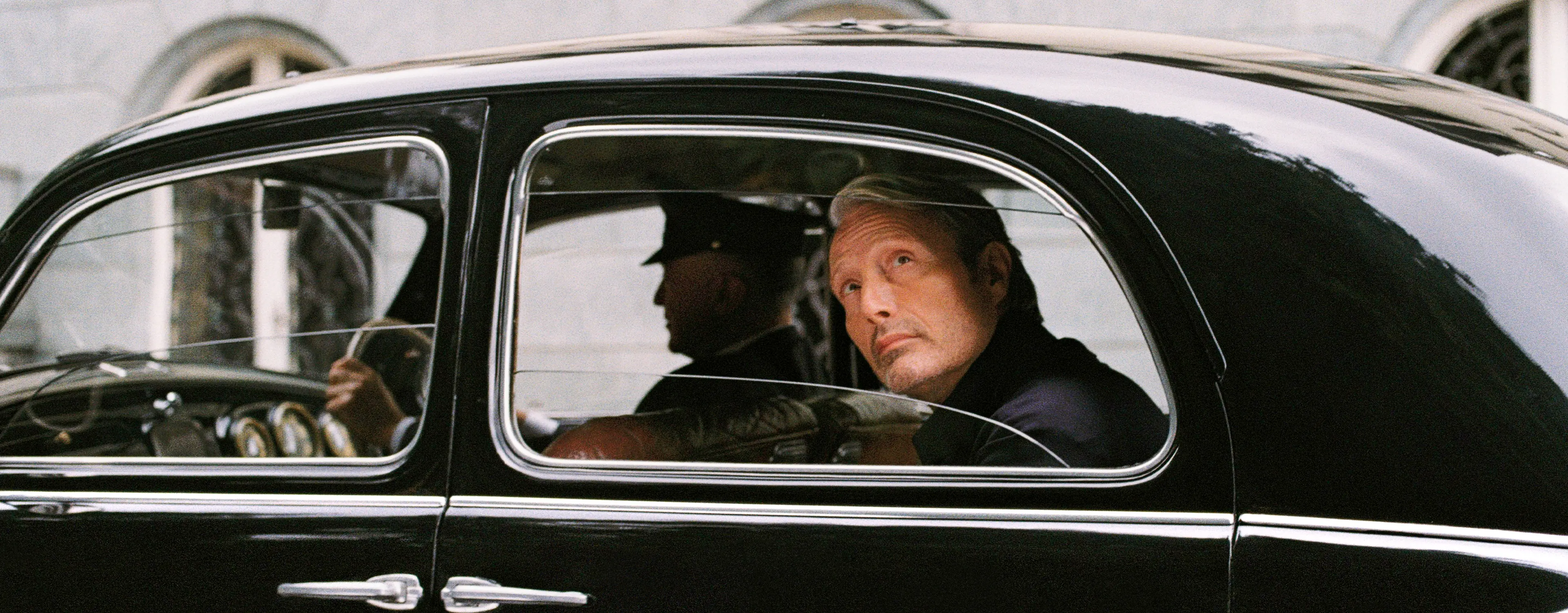 Mads Mikkelsen seated inside a classic black car, looking out through the side window.