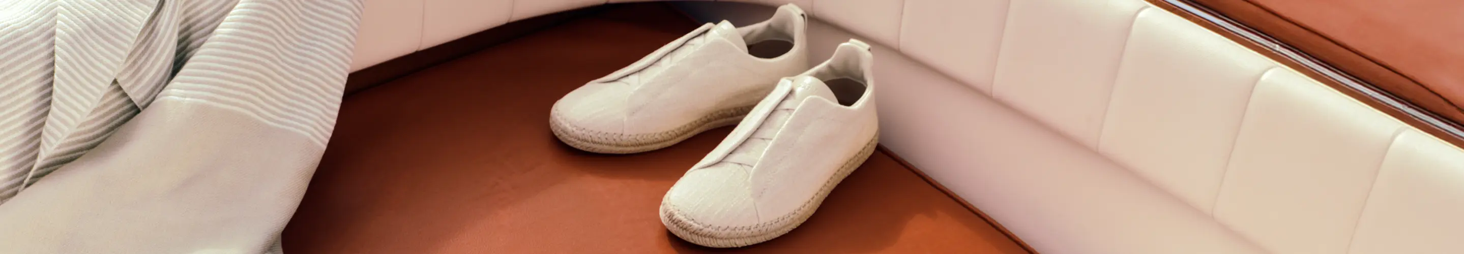 Un par de alpargatas de lino Triple Stitch™ y una toalla de playa de ZEGNA, de la colección Primavera-Verano 2026, con un barco de fondo.